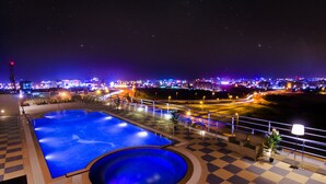 A nighttime view of a pool area with a checkered floor, a large pool, and a cityscape in the background. - Al Murooj Grand Hotel (Muscat)