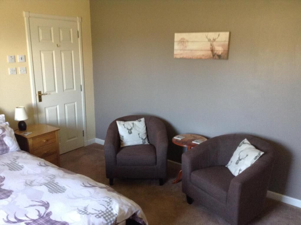 Een slaapkamer met twee fauteuils, een bed, een nachtkastje, een lamp en een schilderij aan de muur.