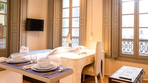 Habitación de hotel con zona de comedor, cama, televisión y ventanas amplias.