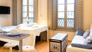 Una habitación de hotel con una cama, una mesa con una silla, una mesita con un reloj, un sofá y ventanas amplias con vistas.