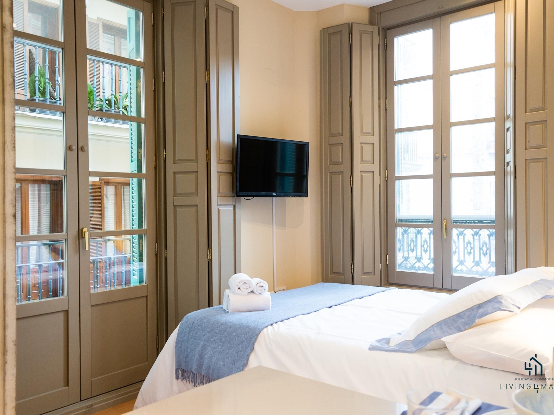A hotel room with a bed, a TV, and large windows with balconies.