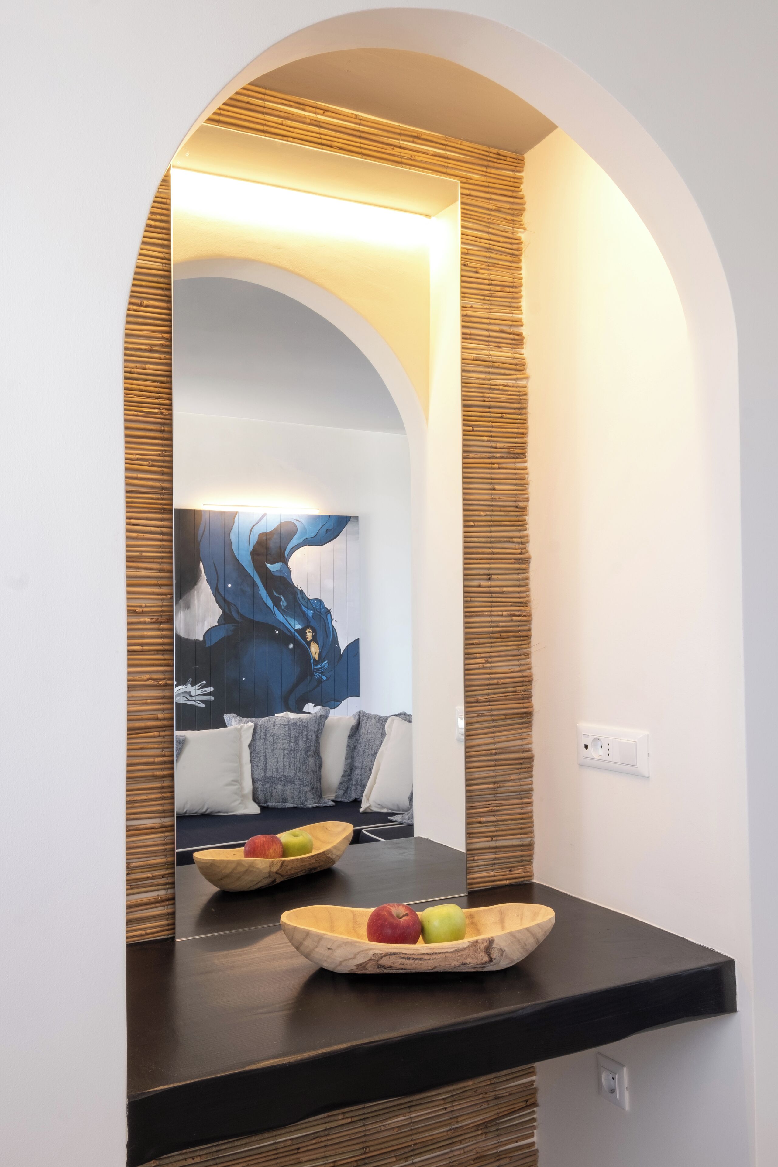 Un interior moderno con una encimera oscura, paneles de madera y un espejo grande que refleja un cuadro y un frutero.