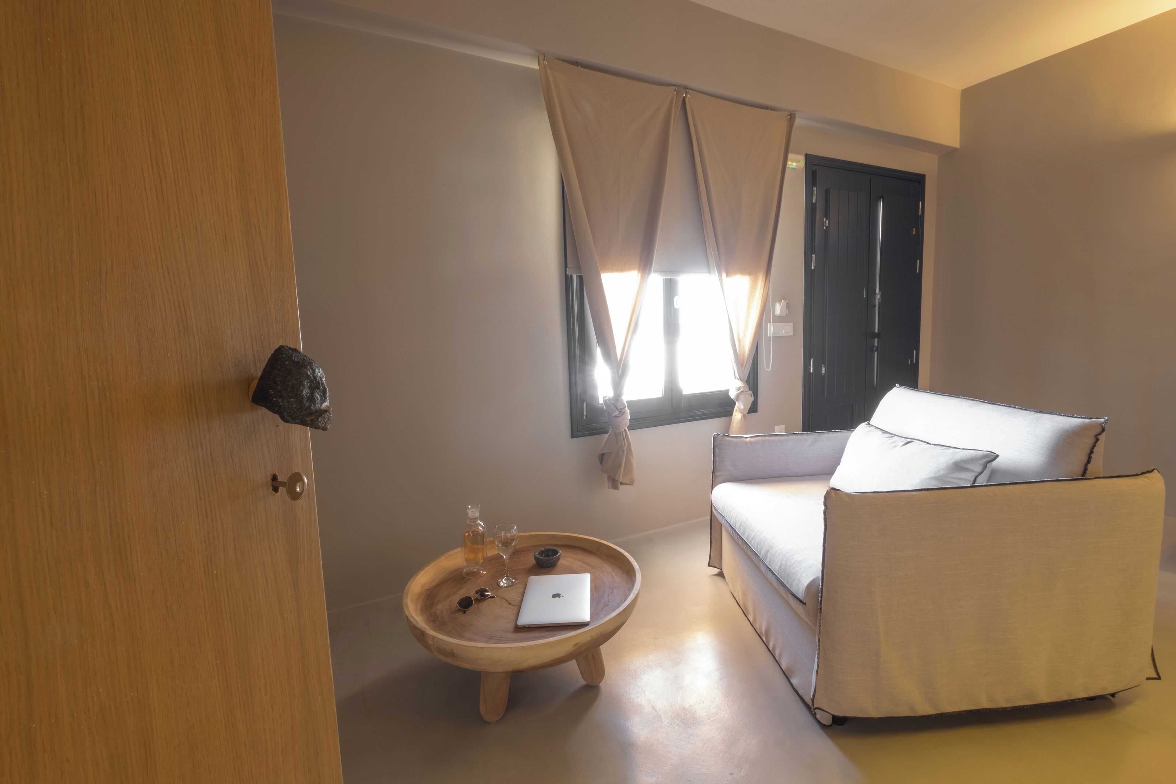 Une pièce avec une porte en bois, une table ronde en bois sur laquelle se trouvent un ordinateur portable, une bouteille et un verre, un canapé beige et une fenêtre avec des rideaux.