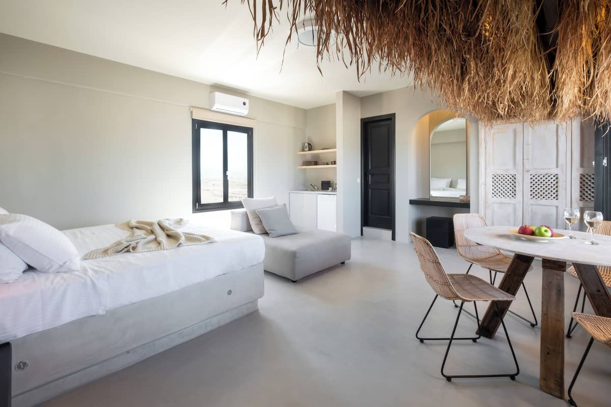 A modern bedroom with a bed, a small sofa, a dining table with chairs, and a kitchen area in the background.