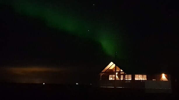 A house with a triangular roof and multiple windows, illuminated by interior lights against a dark sky with green aurora lights. - Black beach cottage (Sveitarfélagid Ölfus)