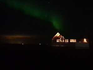 A house with a triangular roof and multiple windows, illuminated by interior lights against a dark sky with green aurora lights. - Black beach cottage (Sveitarfélagid Ölfus)