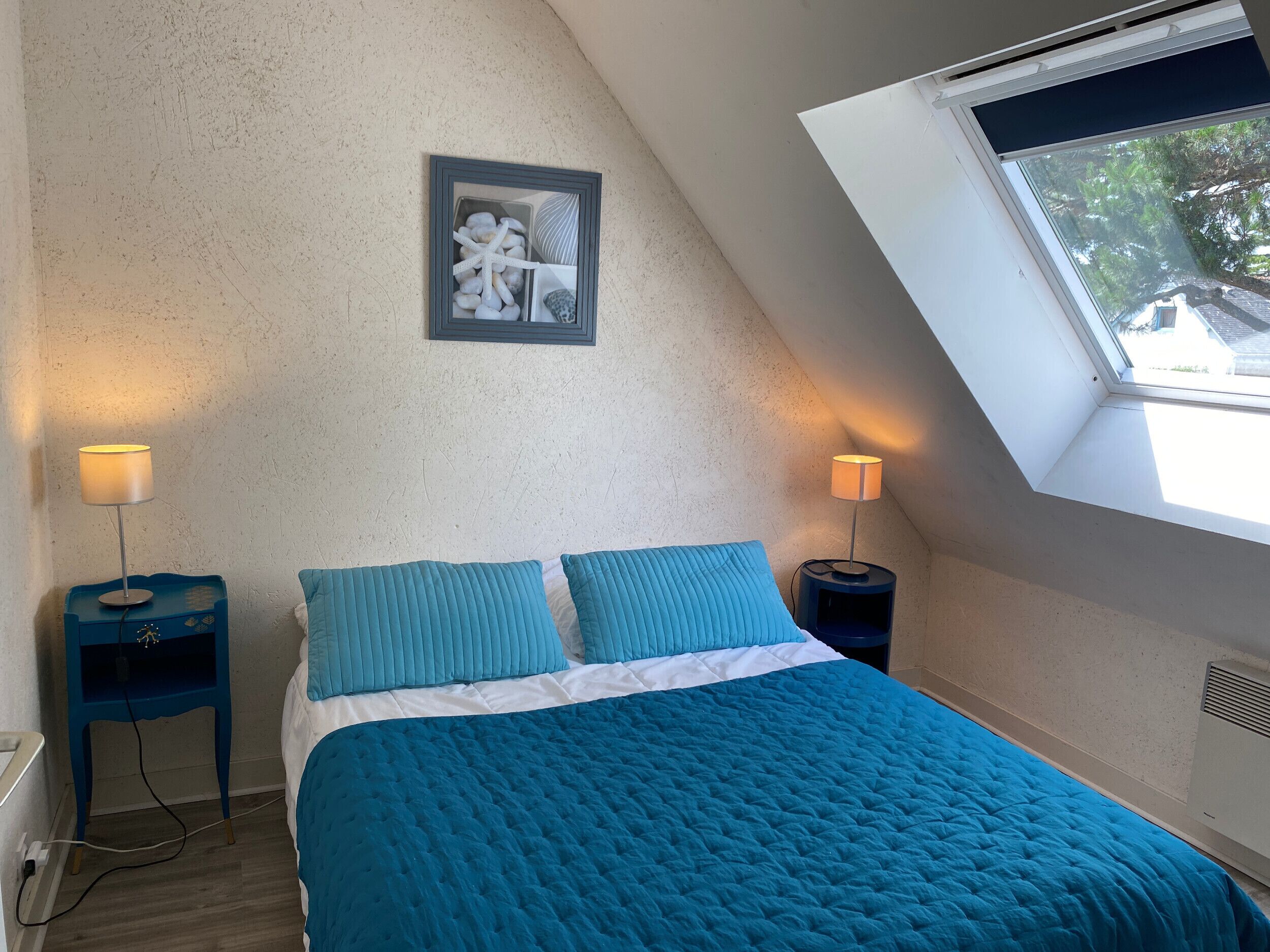 Un dormitorio con techo inclinado, tragaluz, cama con ropa de cama azul, dos lámparas de noche y un cuadro en la pared.