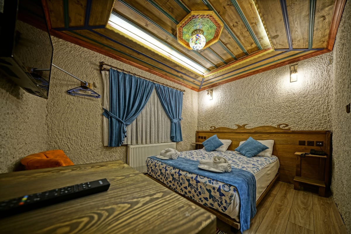 A bedroom with a wooden ceiling, a bed with blue bedding, a nightstand, and a wooden table with a remote control.