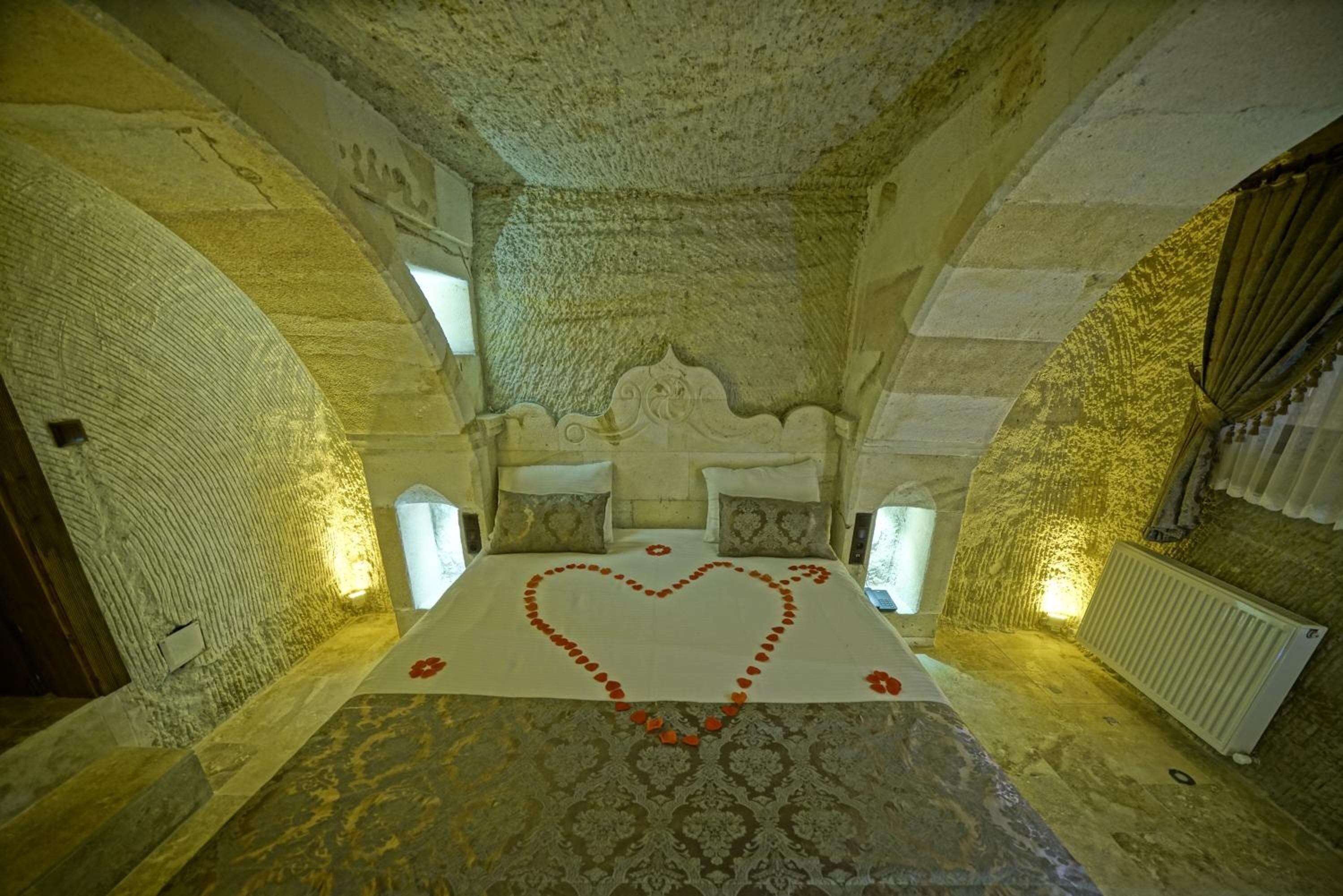 Una habitación con una cama centrada bajo un techo de piedra abovedado, un arreglo de pétalos de rosa en forma de corazón sobre la cama y una alfombra estampada.