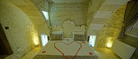 Una habitación con una cama centrada bajo un techo de piedra abovedado, un arreglo de pétalos de rosa en forma de corazón sobre la cama y una alfombra estampada.
