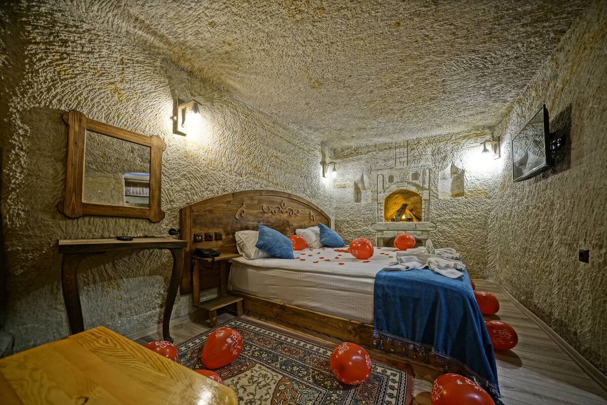 A cozy bedroom with a rustic stone ceiling, a wooden bed with blue bedding, and a small table with a mirror.