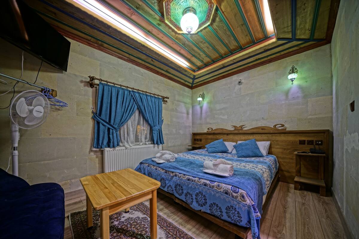 A bedroom with a wooden bed, blue bedding, a wooden nightstand, a bedside table, a wall-mounted fan, and a ceiling light fixture.