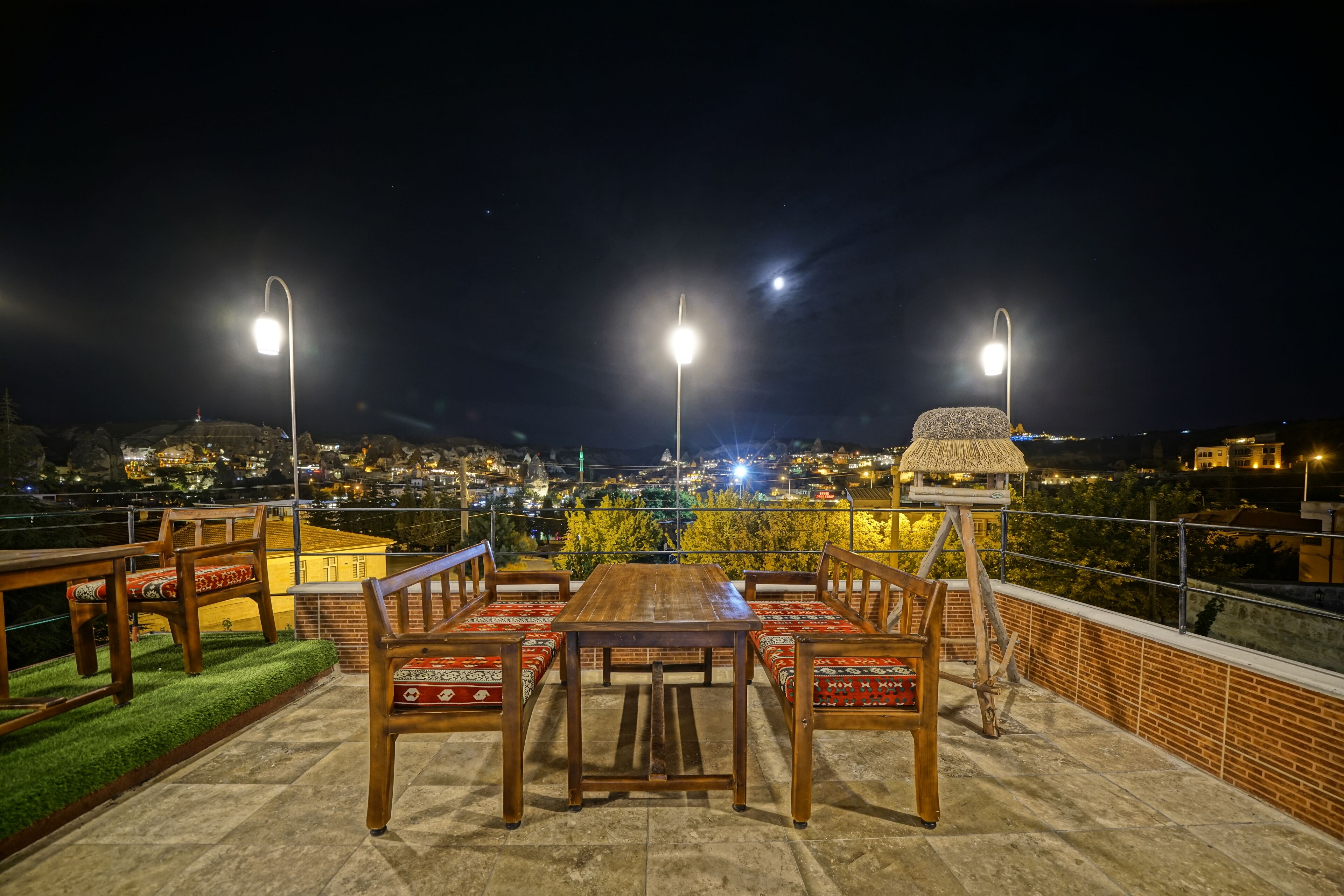 A rooftop terrace with a wooden table and chairs, surrounded by a brick wall and a view of a cityscape at night.