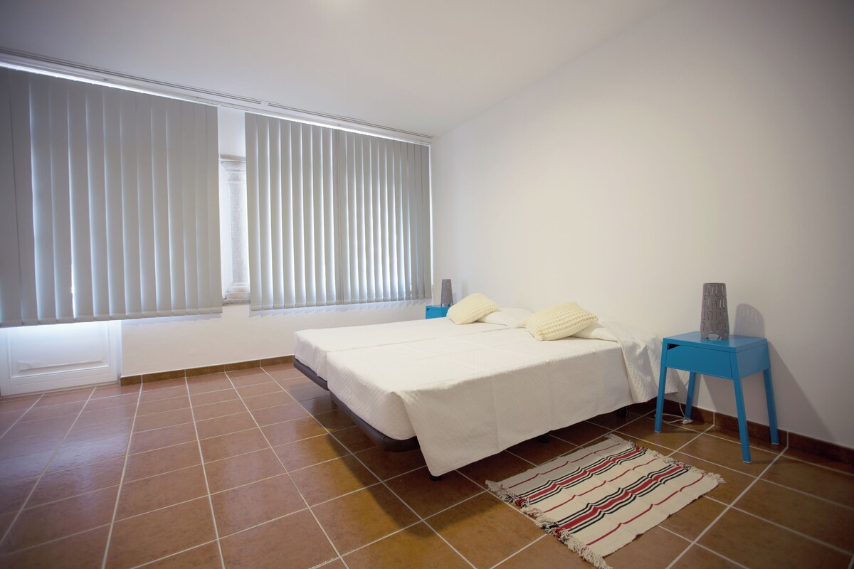 Un dormitorio con dos camas, una mesita de noche azul, una pared blanca y una ventana con persianas.