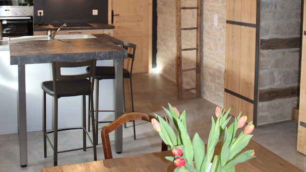A rustic kitchen with exposed beams, a stone wall, and a wooden island with bar stools. - Chambres D'Hôtes Bered Vuillemin (Baume-les-Dames)
