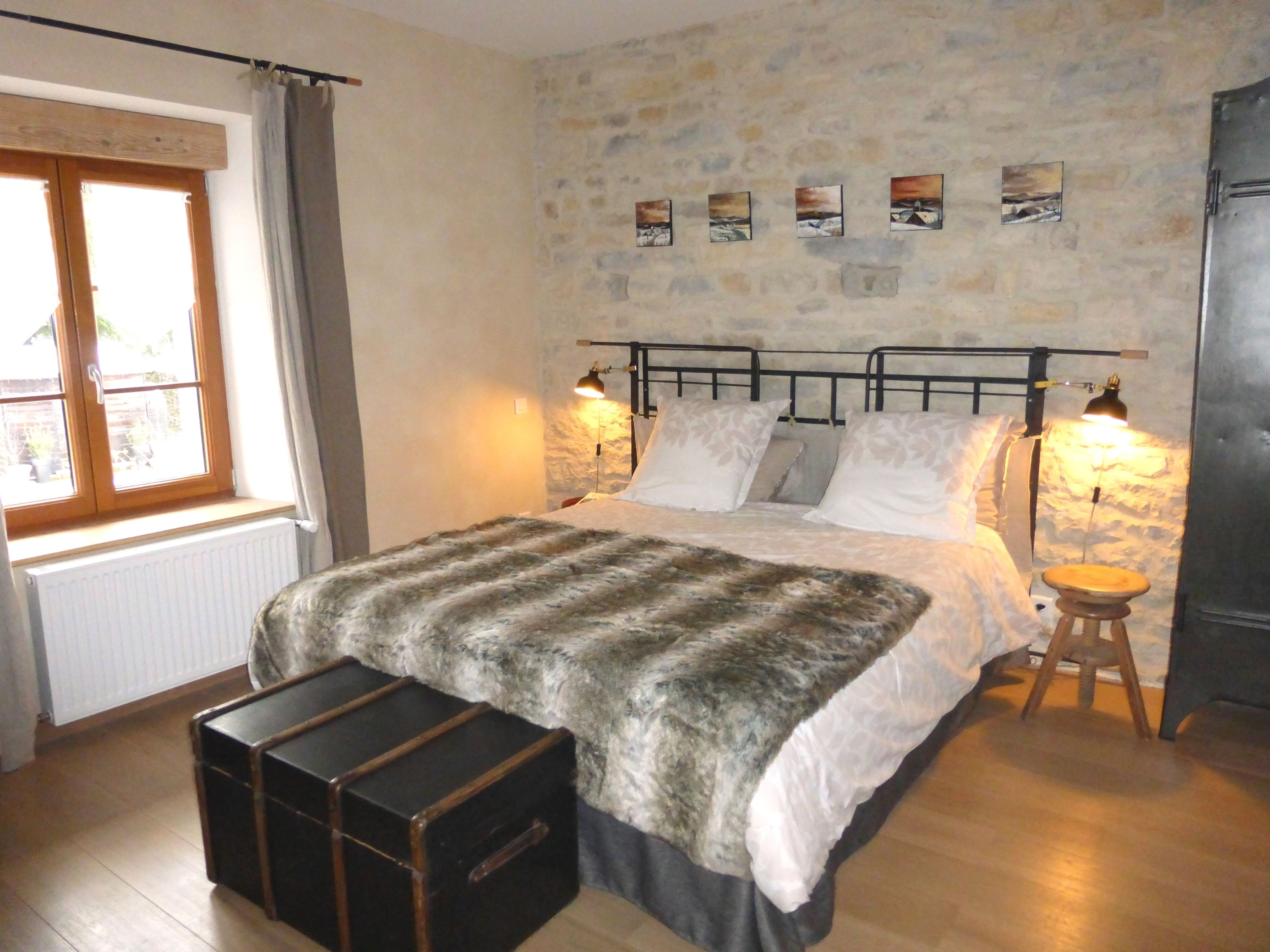 Une chambre avec un mur en pierre, un lit avec une literie blanche, deux lampes de chevet, une fenêtre avec des rideaux et un petit tabouret.