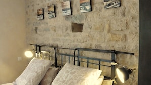 A bedroom with a stone wall, a bed with patterned bedding, a bedside table, and a small stool. - Chambres D'Hôtes Bered Vuillemin (Baume-les-Dames)