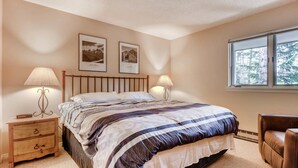 Un dormitorio con cama, dos lámparas de noche, una mesita de noche con cajones, una ventana con vista a árboles y dos cuadros en la pared.