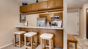 Cocina con armarios de madera, refrigerador de acero inoxidable, microondas y un lavamanos. Hay tres taburetes y un banco.