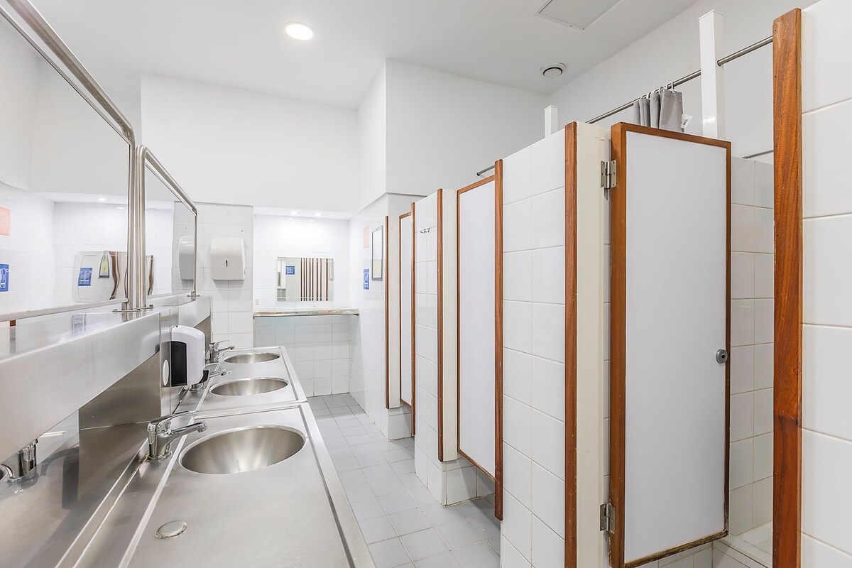 Casa de banho moderna com pias de aço inoxidável, divisórias com estrutura de madeira e chão em azulejo branco.