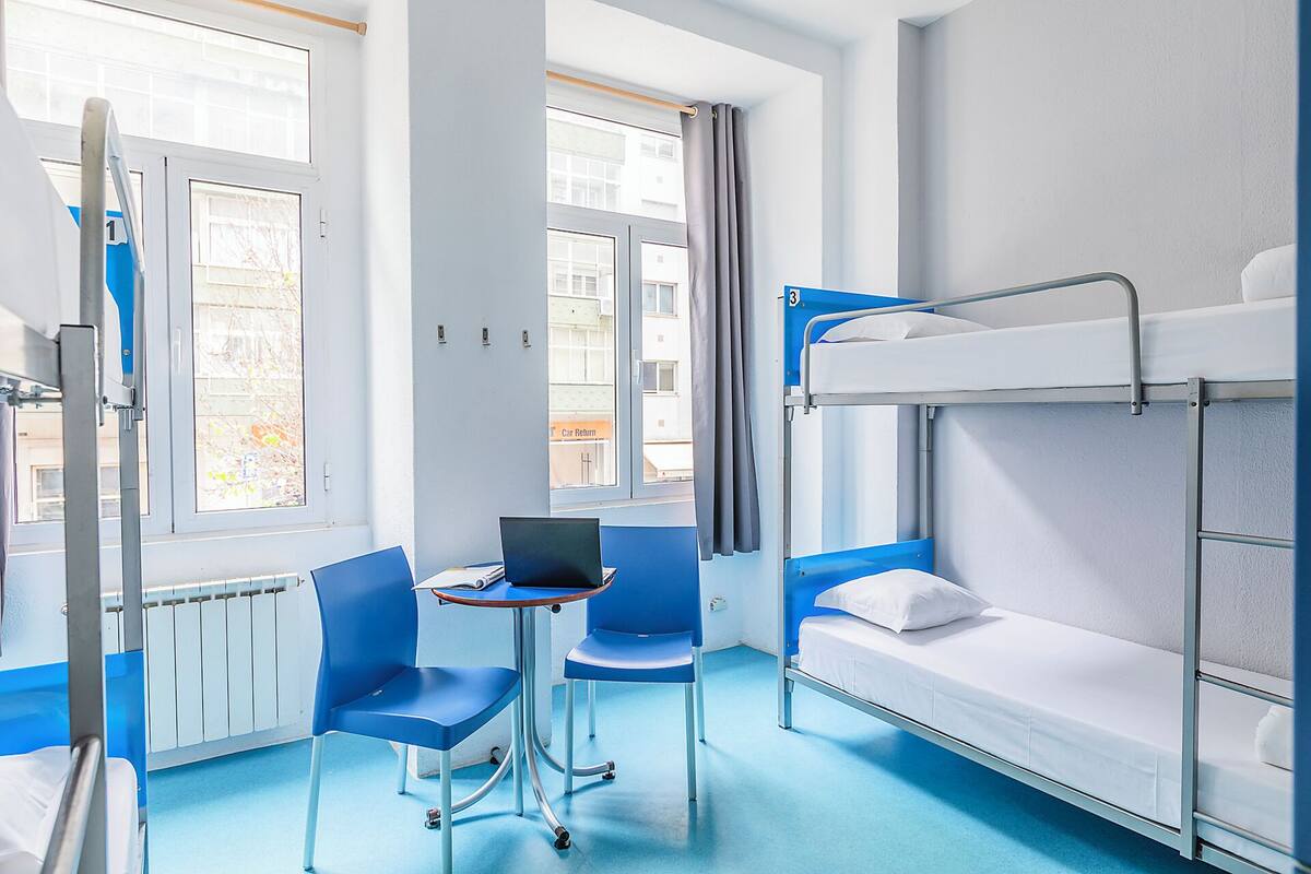 A bunk room with a laptop on a table, two chairs, and a window with curtains.