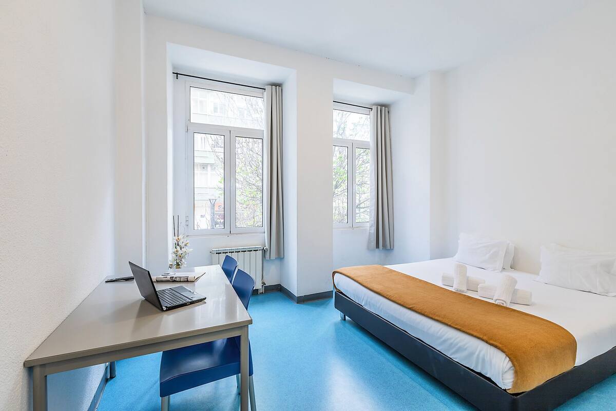 A hotel room with a bed, a desk with a laptop, two windows, and a radiator.