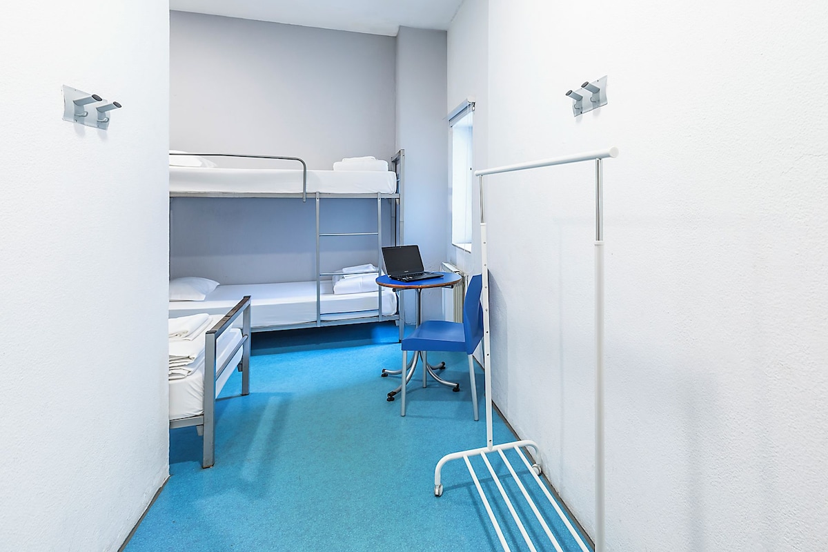 A narrow hotel room with bunk beds, a small desk with a laptop, and a blue chair.