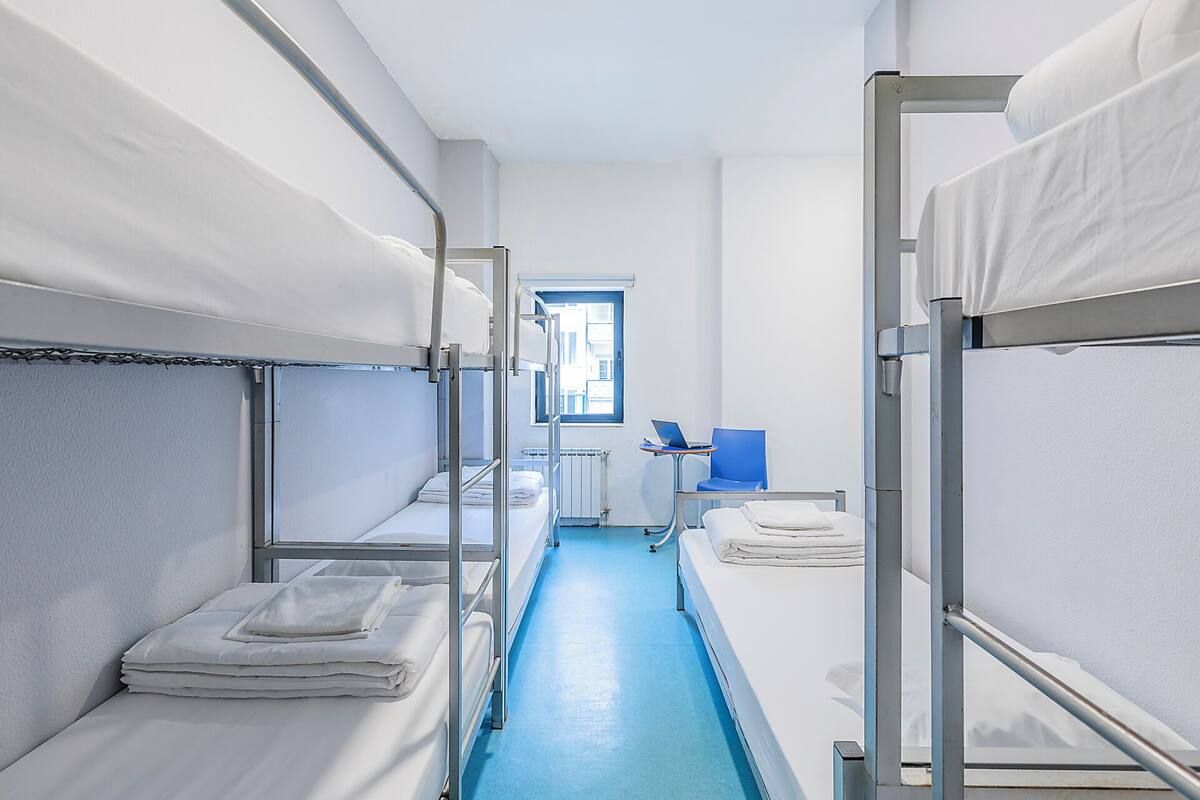 A narrow corridor with bunk beds on either side, a window with a view of buildings, and a small table with a laptop.