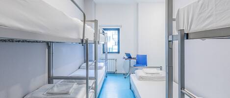 A narrow corridor with bunk beds on either side, a window with a view of buildings, and a small table with a laptop.