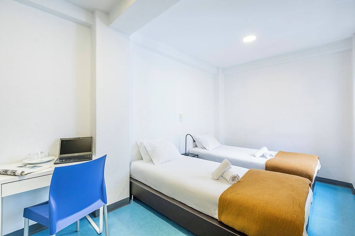 A hotel room with a desk, two beds, a chair, and a ceiling light.