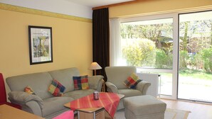 A living room with a sofa, armchair, ottoman, and a small table with a lamp. - Apartmenthaus Padua (Büsum)