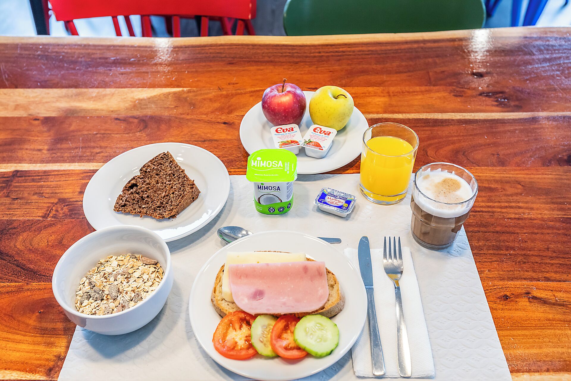 A breakfast setting with a plate of toast, ham, tomatoes, cucumbers, a bowl of cereal, a plate with apples and a glass of orange juice, a cup of coffee, and a container of Mimosa.
