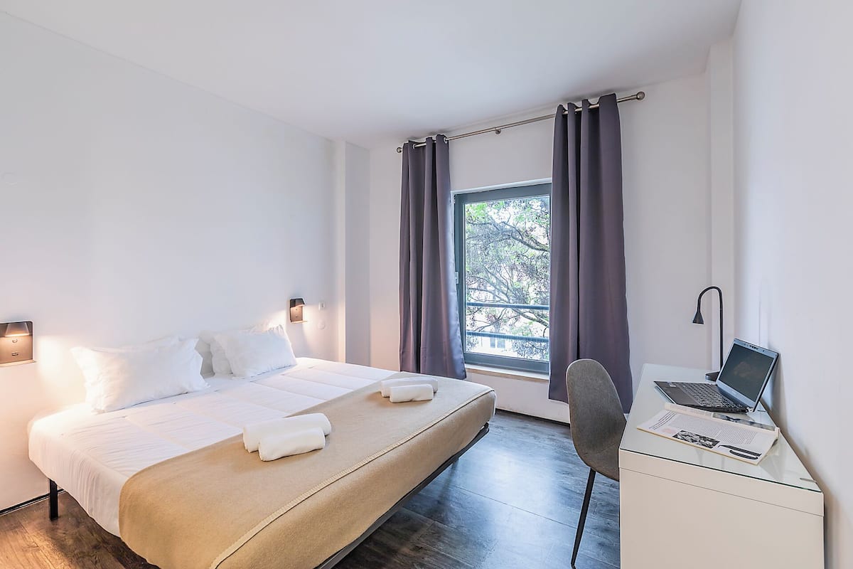 Ein Hotelzimmer mit einem Bett, einem Schreibtisch mit einem Laptop, einem Sessel und einem Fenster mit Vorhängen.
