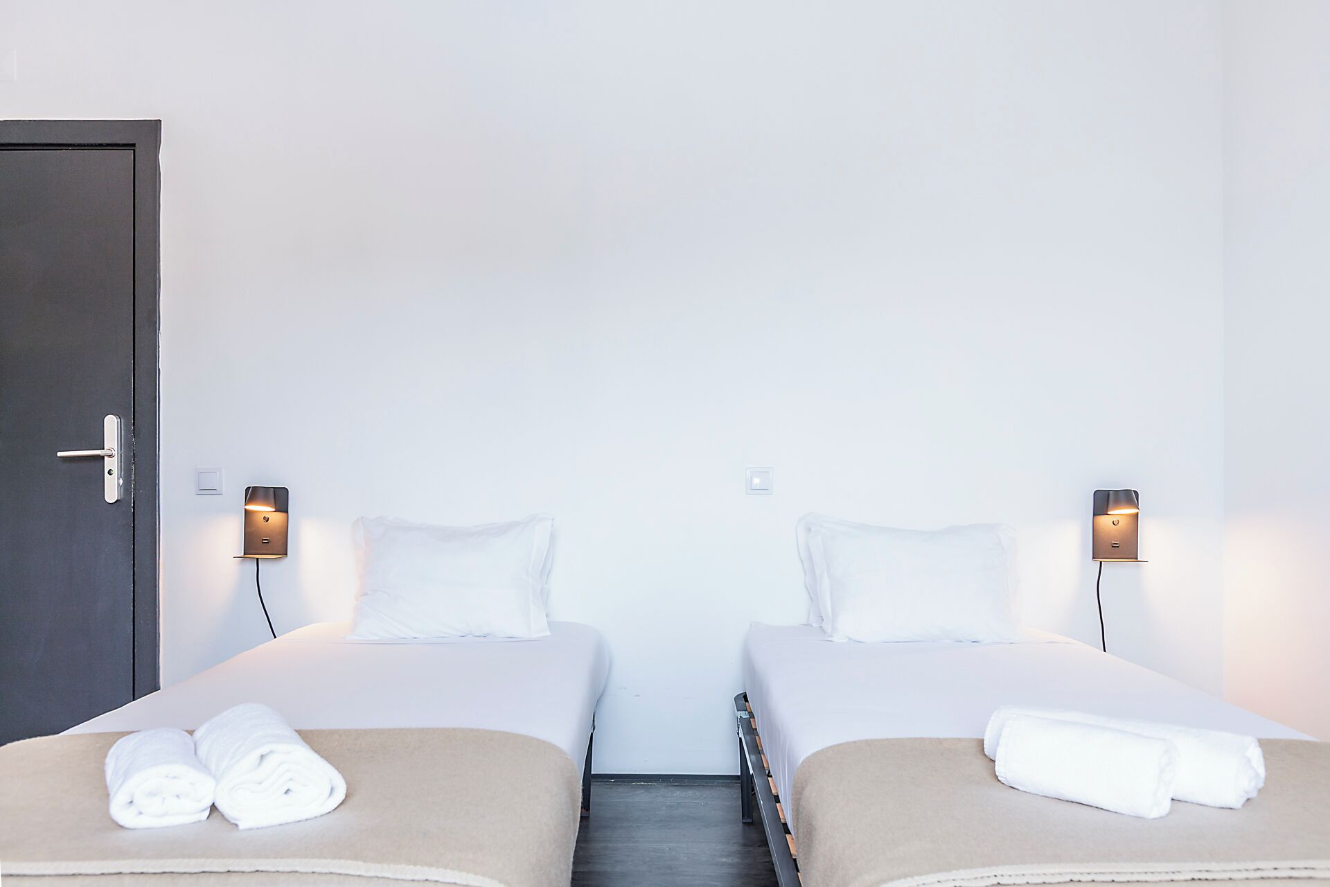 A hotel room with two beds, a door, and wall-mounted light fixtures.