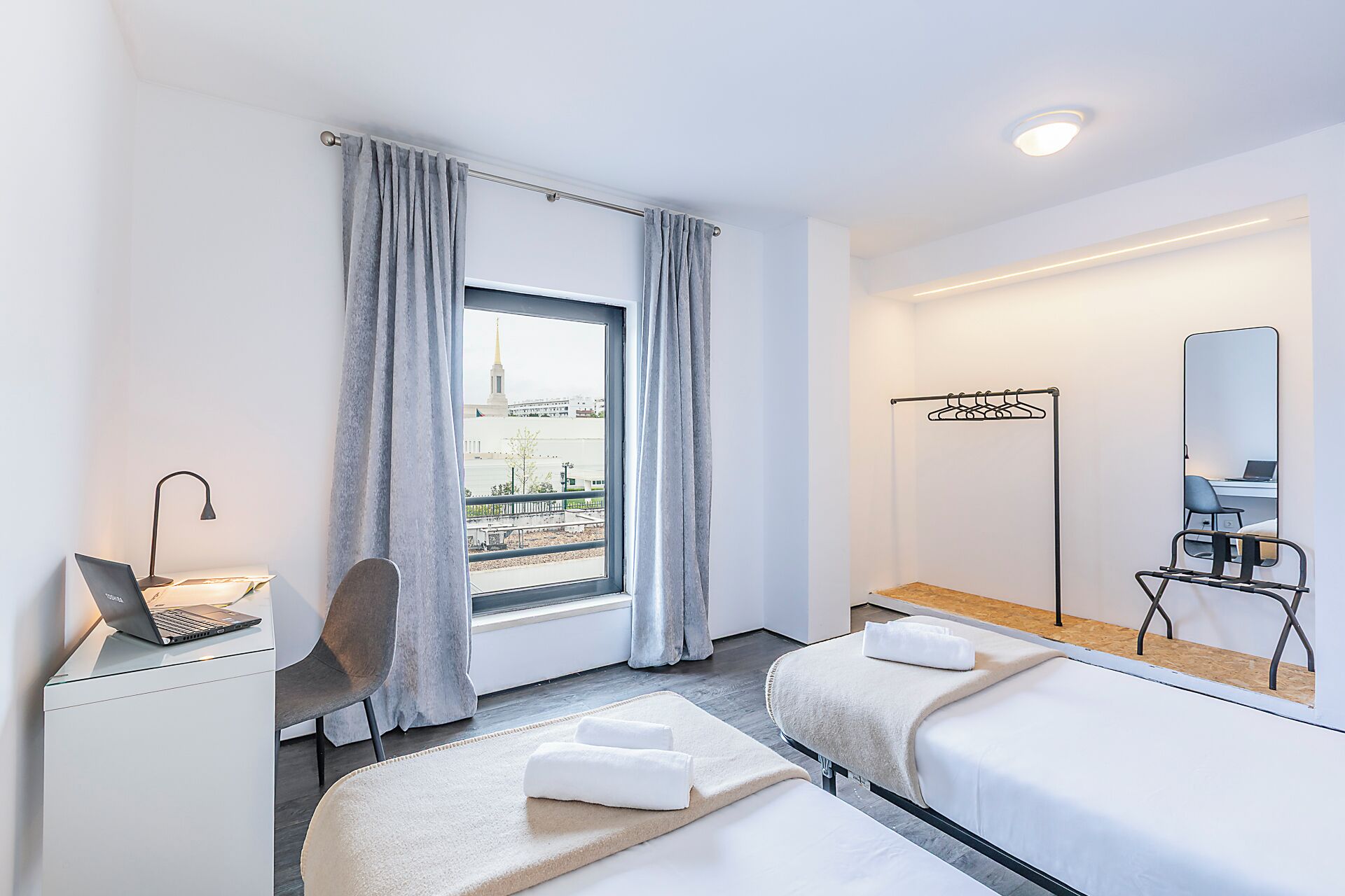 Hotellihuone, jossa on sänky, työpöytä kannettavalla tietokoneella, tuoli, ikkuna verhoilla, peili ja naulakko.