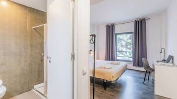 Quarto de hotel moderno com chuveiro, cama com toalhas, escrivaninha com laptop e janela com cortinas.