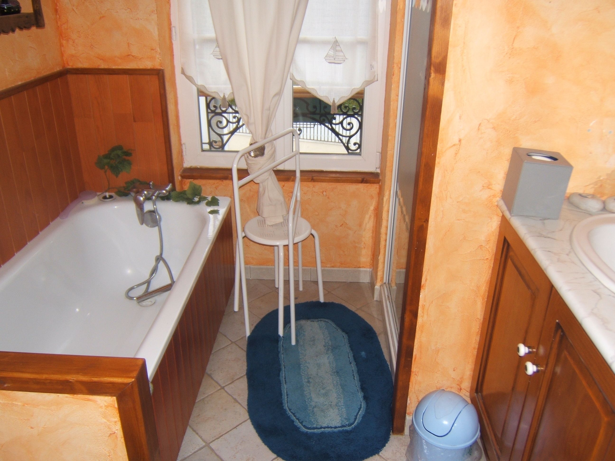 A bathroom with a bathtub, a white sink, a wooden cabinet, a blue rug, and a window with a curtain.