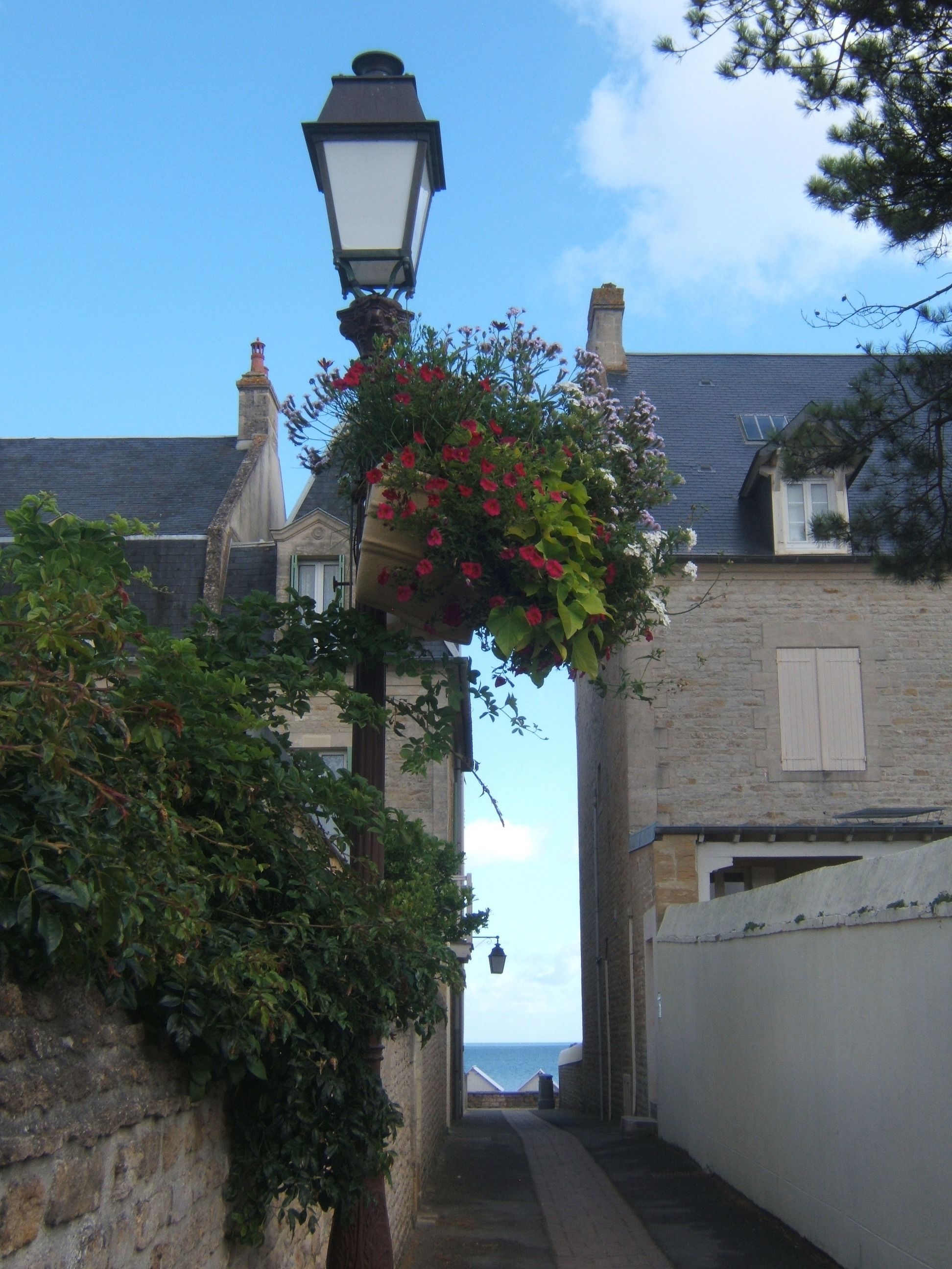 A narrow street with a hanging flower basket, stone walls, and a lamp post.