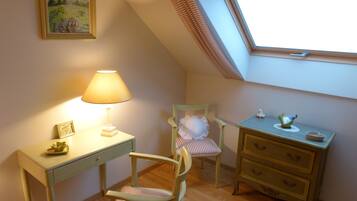 A cozy room with a desk, chair, bedside table, and a window with blinds.