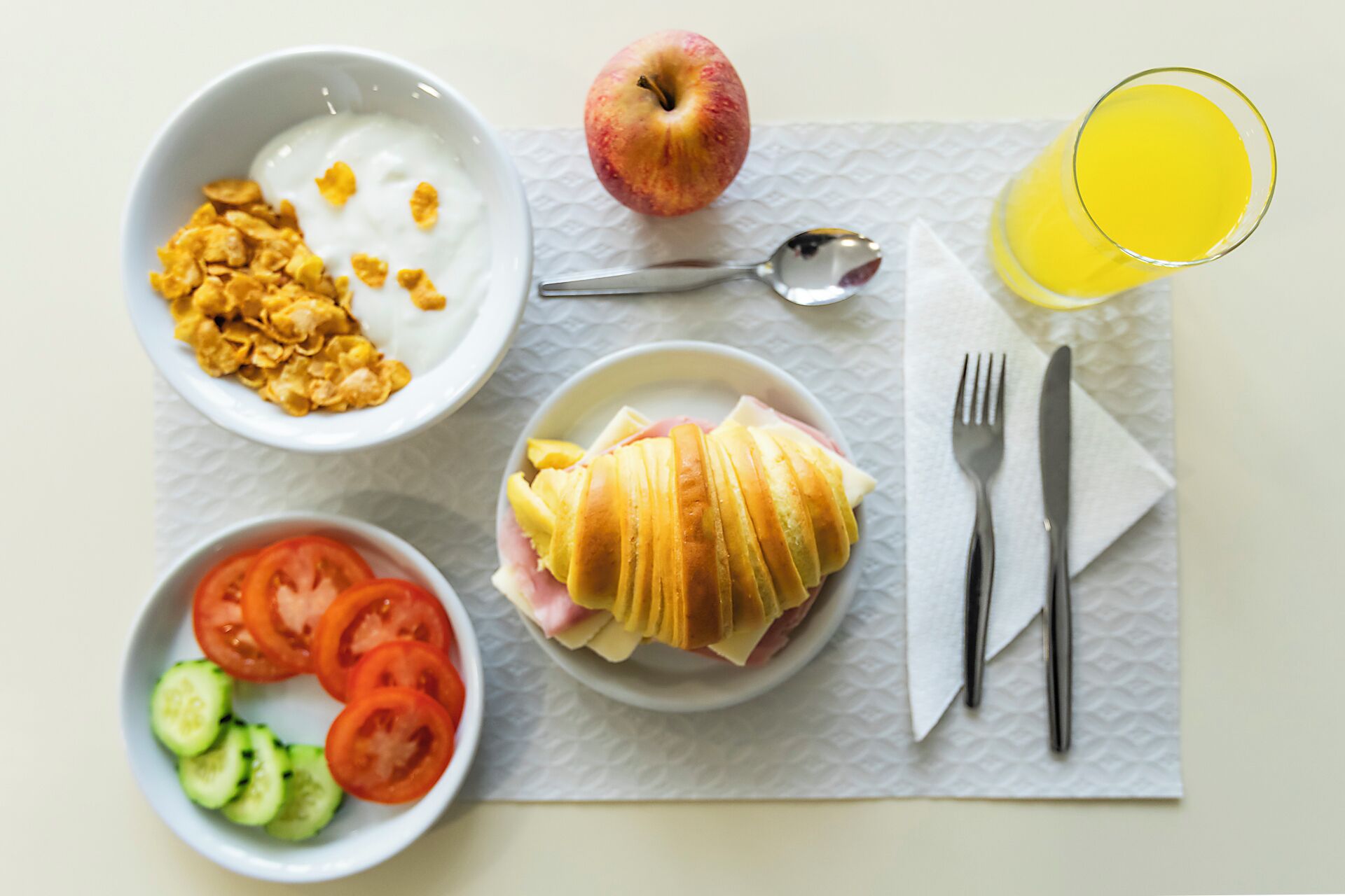 Uma mesa de café da manhã com uma tigela de cereal, um copo de suco de laranja, um sanduíche e um prato com rodelas de tomate e pepino.