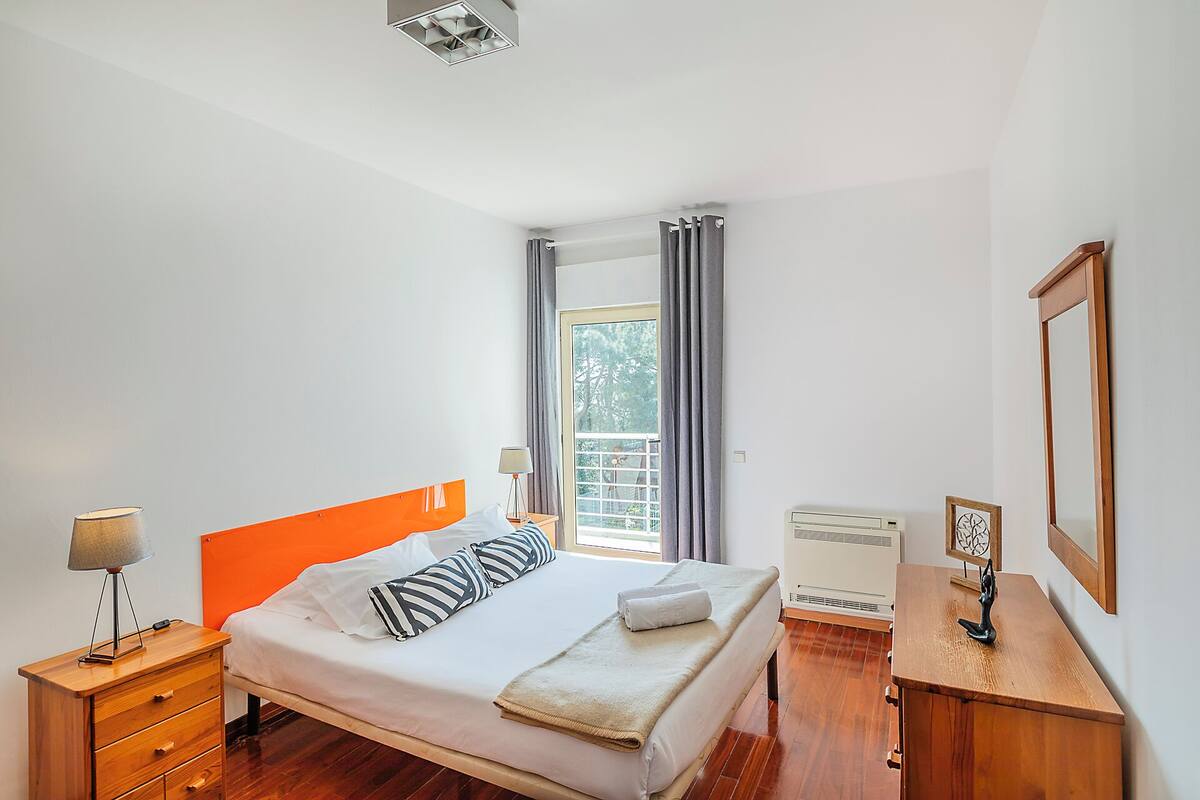 Une chambre d’hôtel avec un lit, une table de chevet en bois, une lampe, une fenêtre avec des rideaux, un radiateur et un tableau encadré au mur.