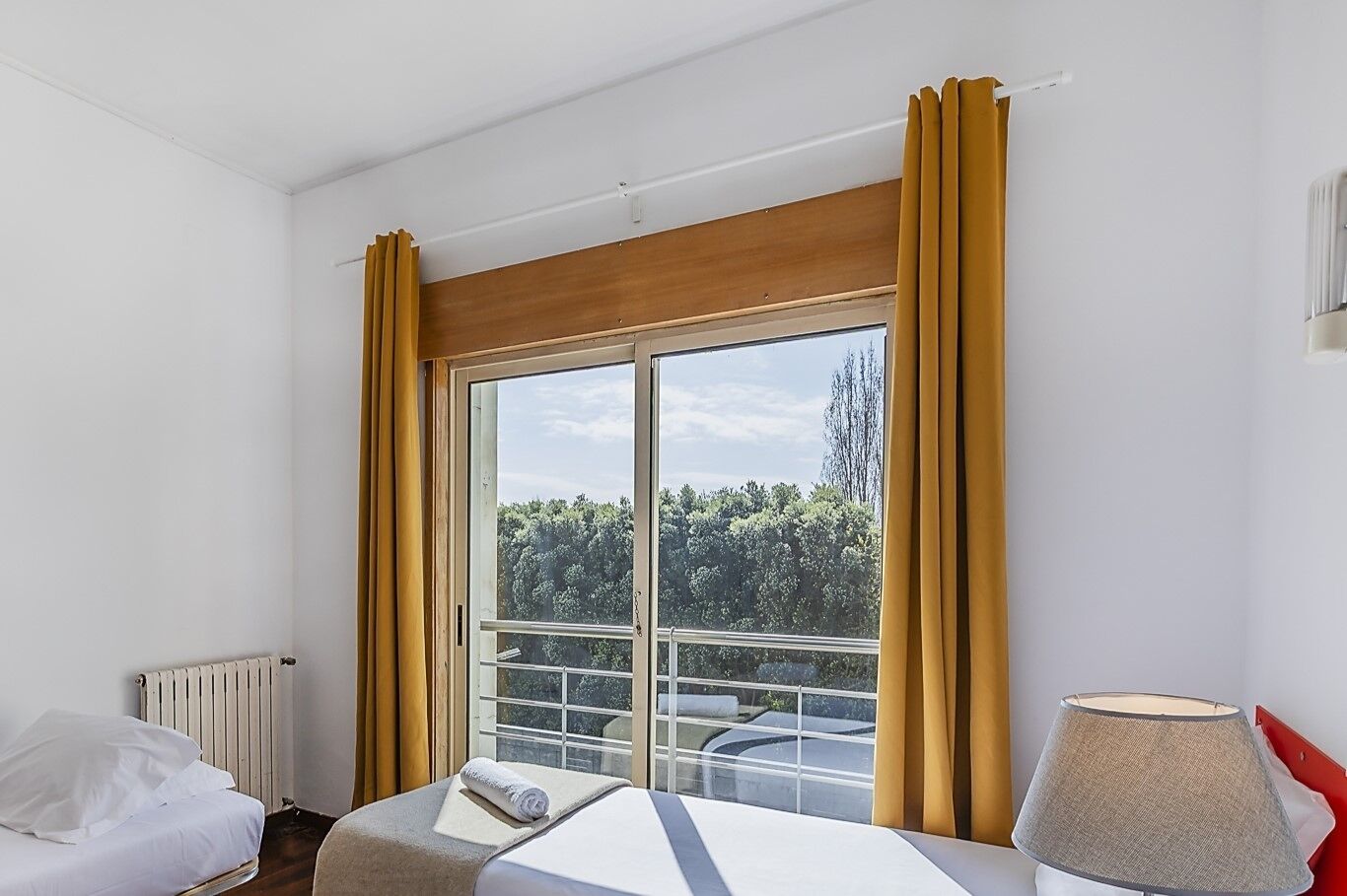 A hotel room with a bed, a window with curtains, a radiator, and a lamp.