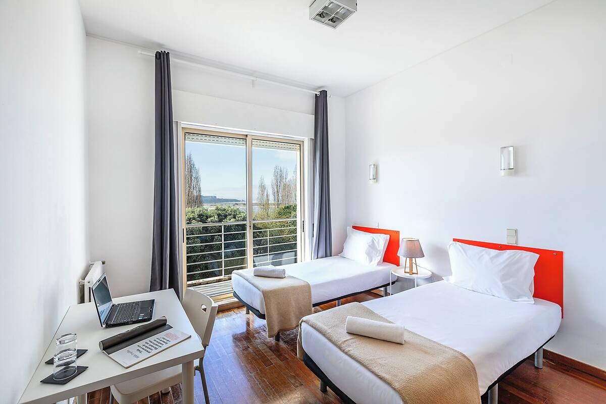 Une chambre d’hôtel avec deux lits, un bureau avec un ordinateur portable, une chaise et une vue sur la verdure environnante.