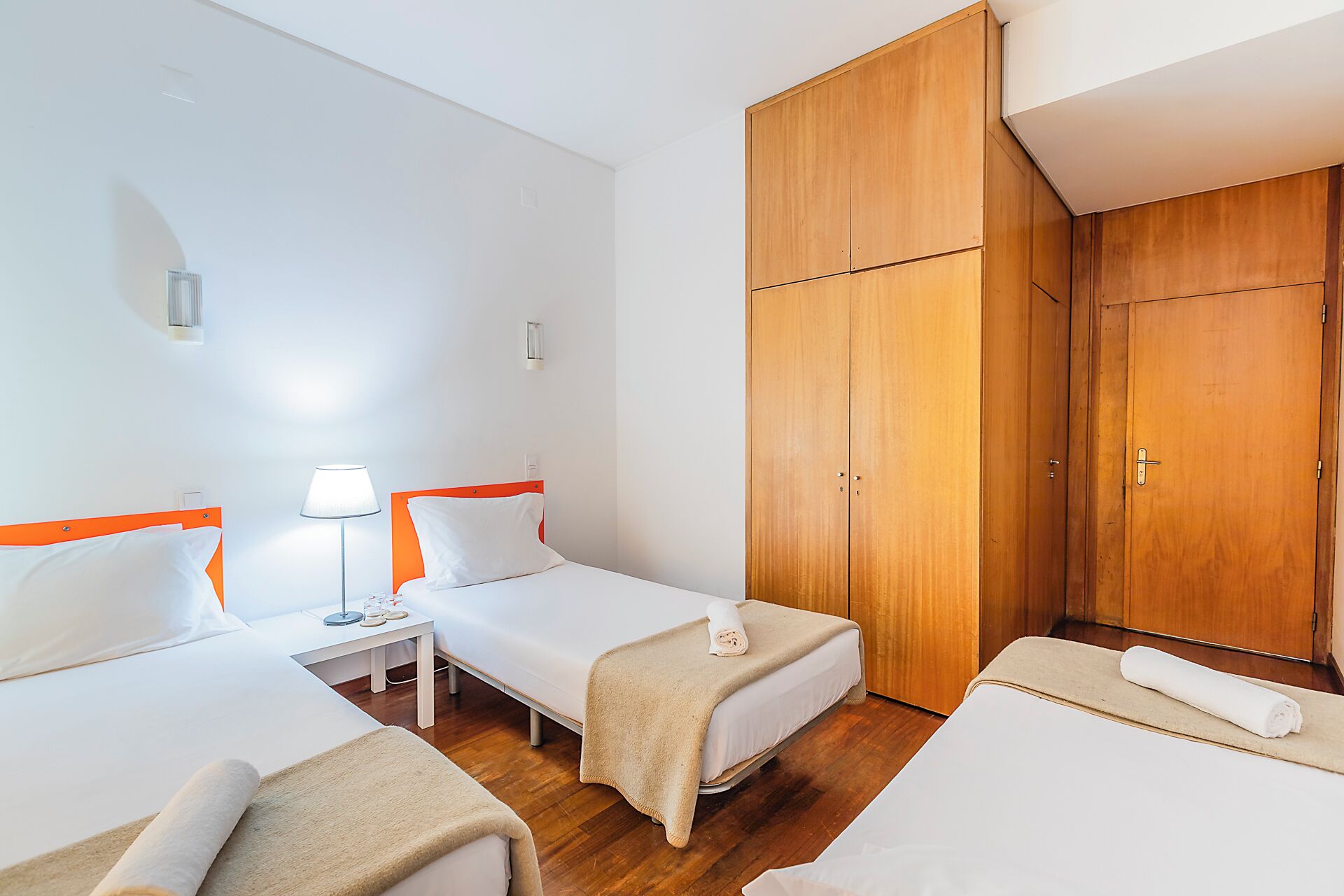 Habitación de hotel con dos camas individuales, un armario de madera, una mesita de noche con lámpara y una pequeña mesita auxiliar.