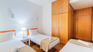 Habitación de hotel con dos camas individuales, un armario de madera, una mesita de noche con lámpara y una pequeña mesita auxiliar.