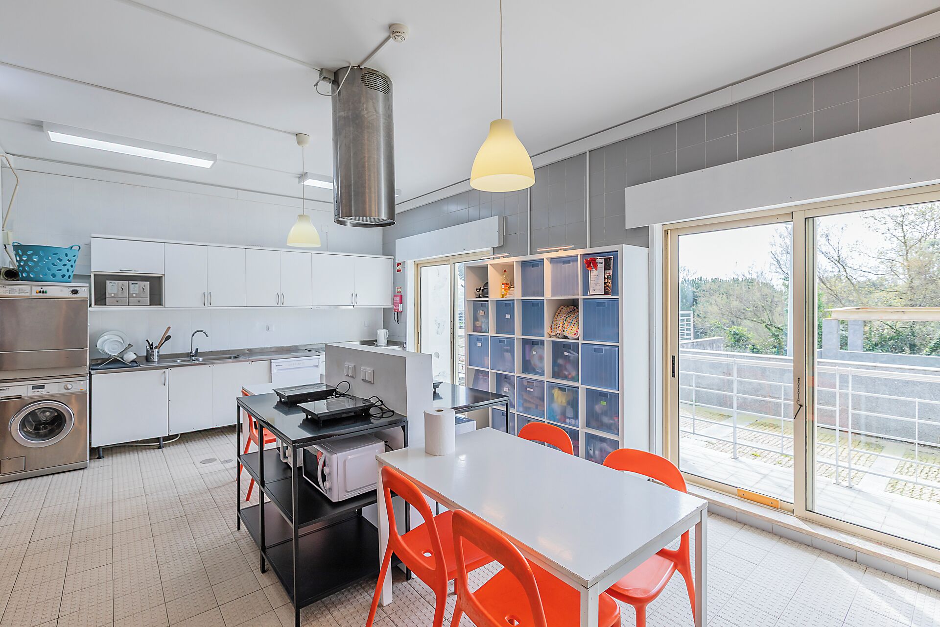 A modern kitchen with stainless steel appliances, a dining area with orange chairs, and a large window offering a view of the outdoors.