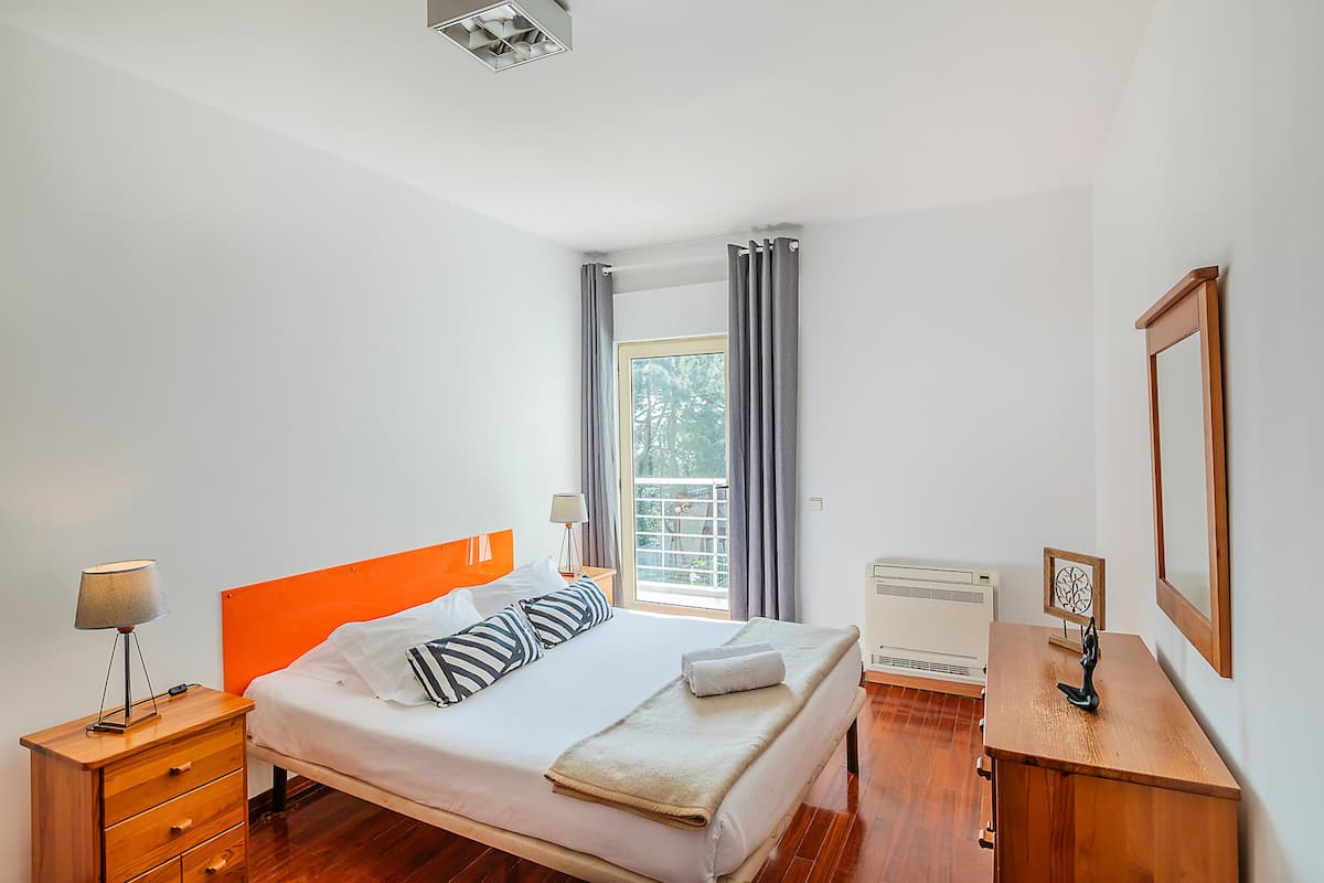 Une chambre à coucher avec un lit, des tables de chevet, une fenêtre avec des rideaux, un radiateur et un miroir au mur.