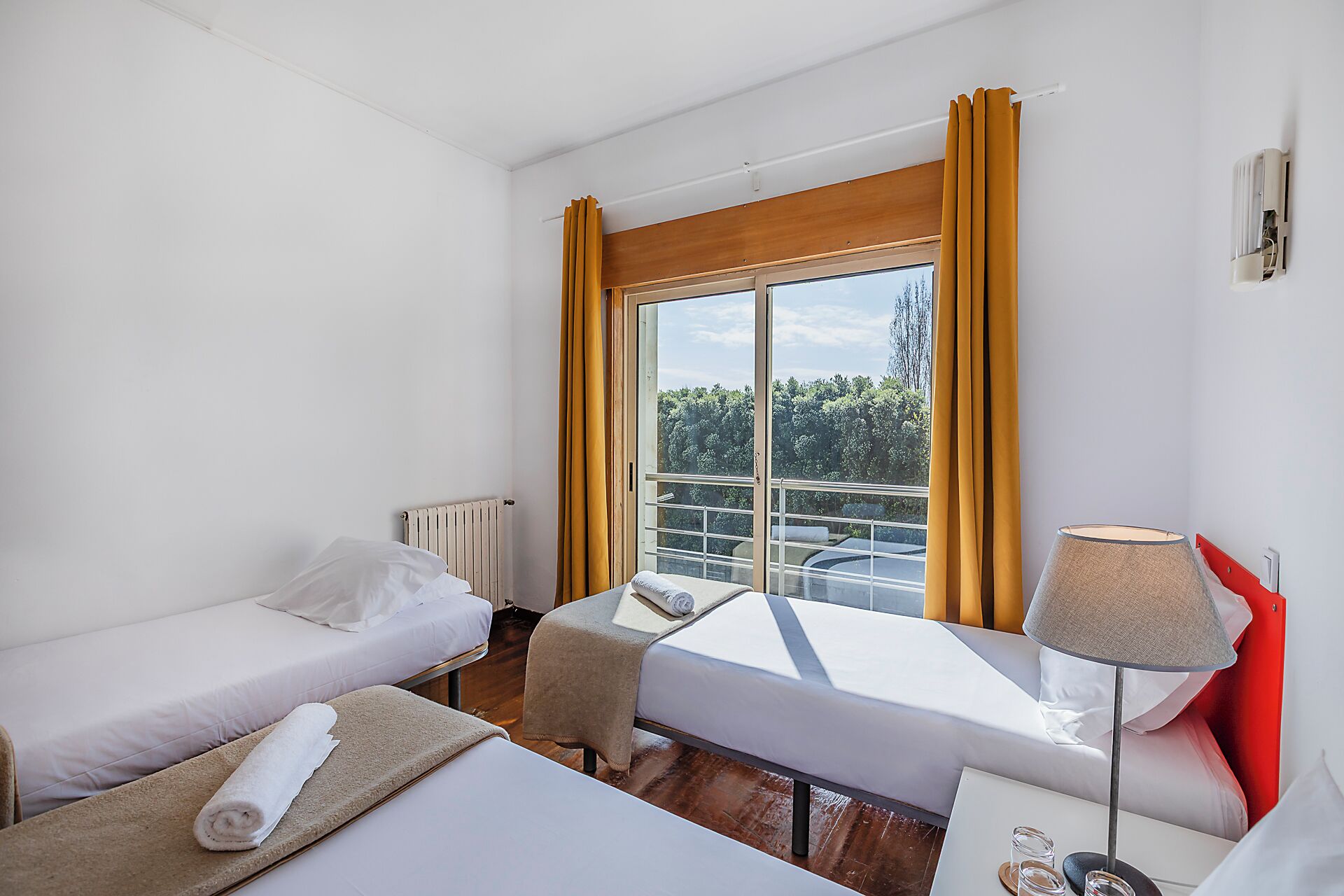 Quarto de hotel com duas camas, uma janela com cortinas, um radiador, uma mesa com abajur e vista para o verde lá fora.