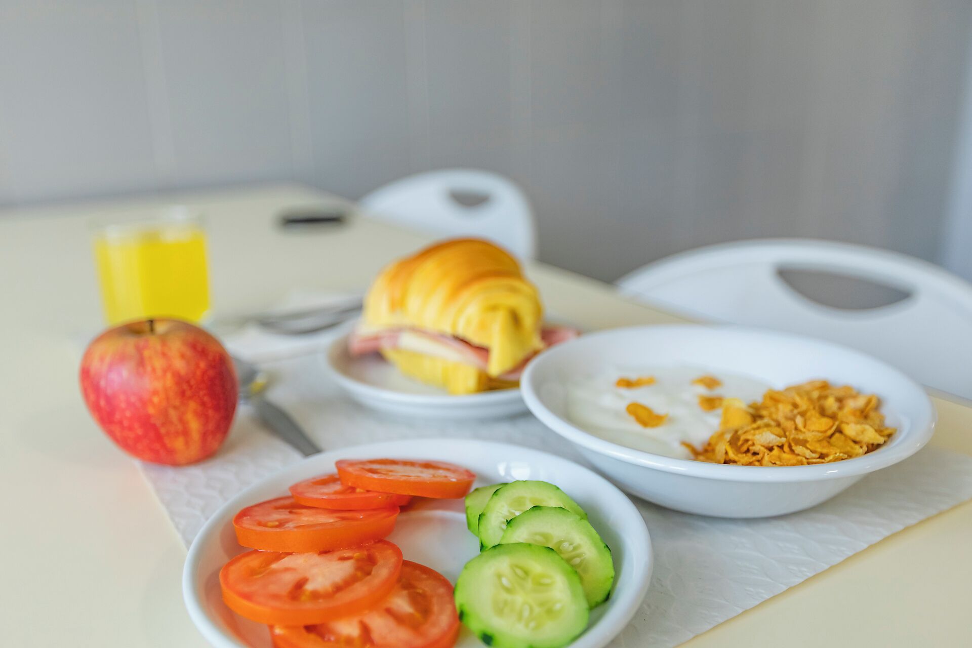 A breakfast table with a croissant, apple, tomato slices, cucumber slices, yogurt, and cereal.