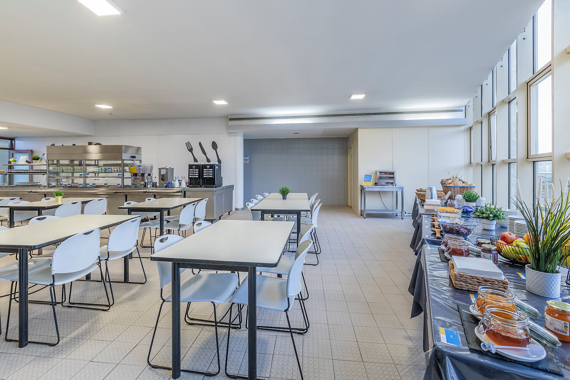 A cafeteria with tables and chairs, a kitchen area, and a buffet table with food items.